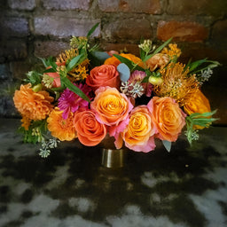 orange roses, ranunculus, protea, eucalyptus in a gold vase