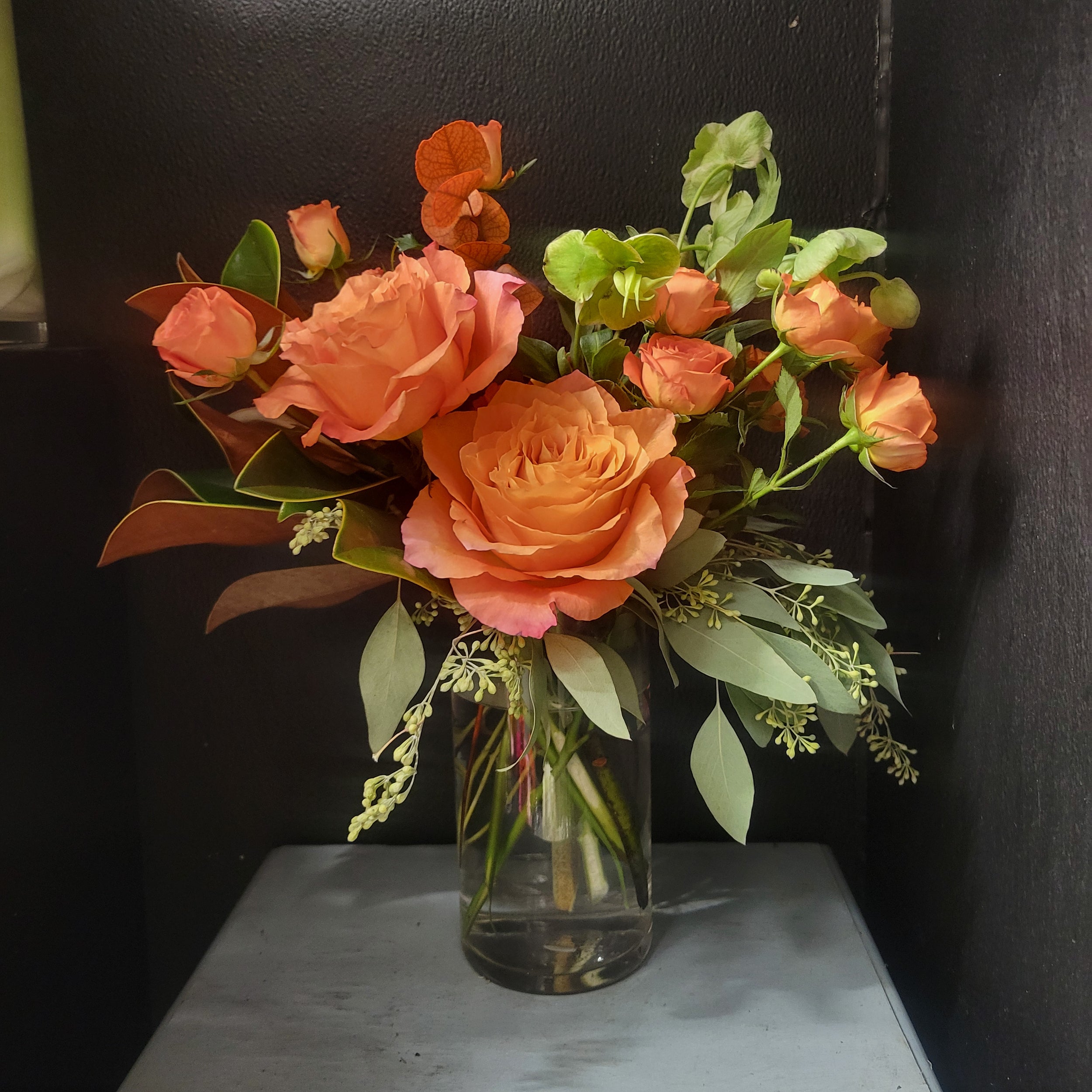 Bouquet of orange roses and greenery in a clear vase on a dark surface.